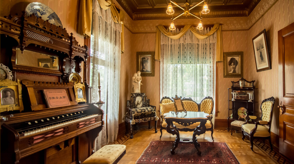 The front parlor at Rosson House, with electric lighting, light pink wallpaper, a gold pressed tin ceiling, parquet wood flooring, and ornate decorations.