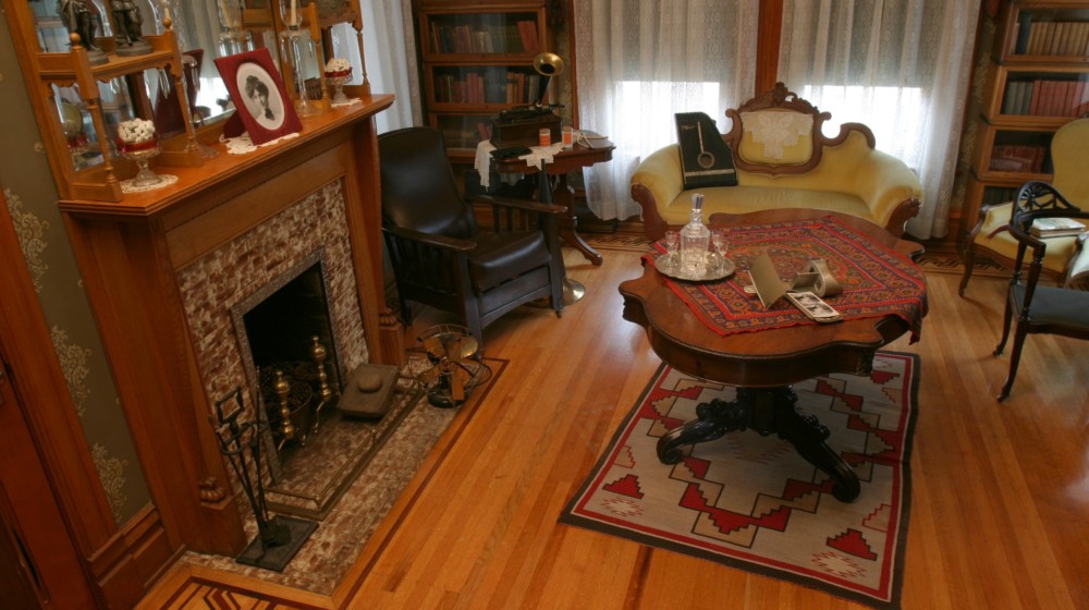 The back parlor at Rosson House with sage green wallpaper, parquet wood flooring, a fireplace, two tall bookcases, a phonograph, and other period antiques.