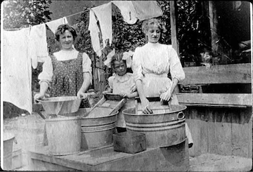 A woman with her two daughters, one a teenager and the other around 8 years old, washing laundry in tubs outside their home, circa 1900.