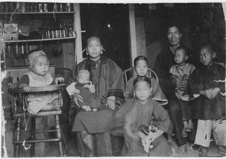 Black and white picture of a Chinese immigrant family from 1893. They are in a small room, and there are two parents and six young children of varying ages.