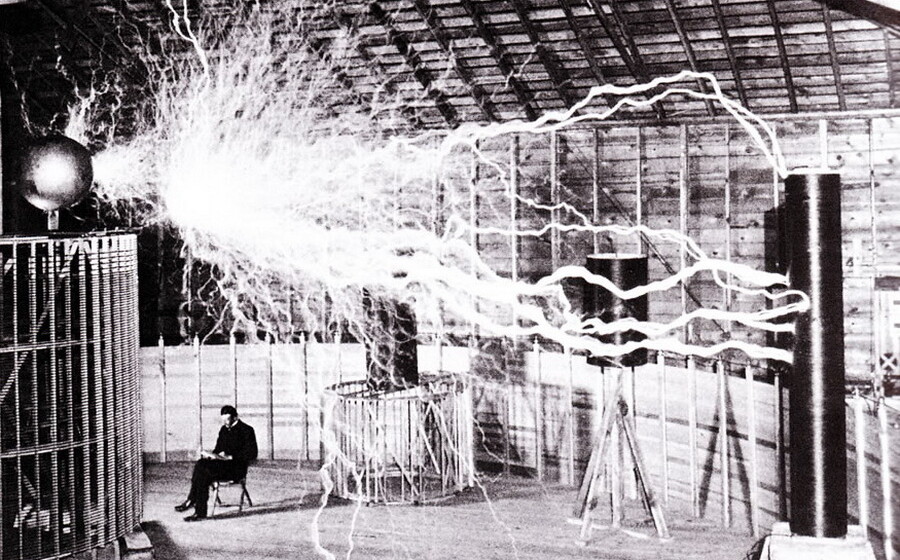 A black and white picture of Nikola Tesla and his invention, the Tesla Coil, with great arcs of electricity from one coil to another.
