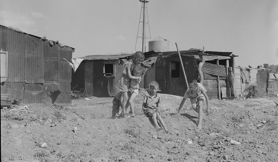 A black and white picture of a small group of kids playing outside shacks at an Arizona migrant camp during the Great Depression, circa May 1937.